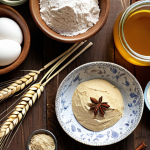 A beautifully arranged table featuring various organic baking ingredients. Include organic wheat flour, *siyez* flour, rye flour, honey, *pekmez*, maple syrup, olive oil, coconut oil, butter, eggs, milk, and spices like cinnamon. The scene should evoke a sense of healthy and delicious homemade treats.  Emphasize natural light and rustic textures.